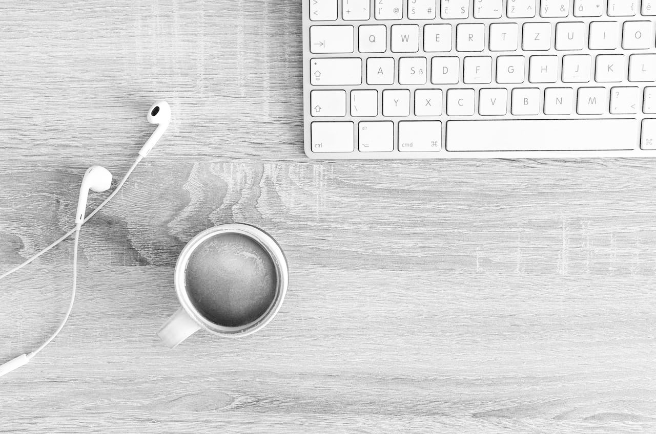 Image of a light colored desk with white keyboard, a cup of coffee and a pair of ear bud headphones.